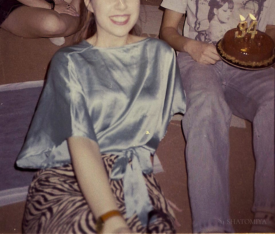 Photo of a girl in clear focus smiling, while behind her a boy holds a birthday cake with lit candles.