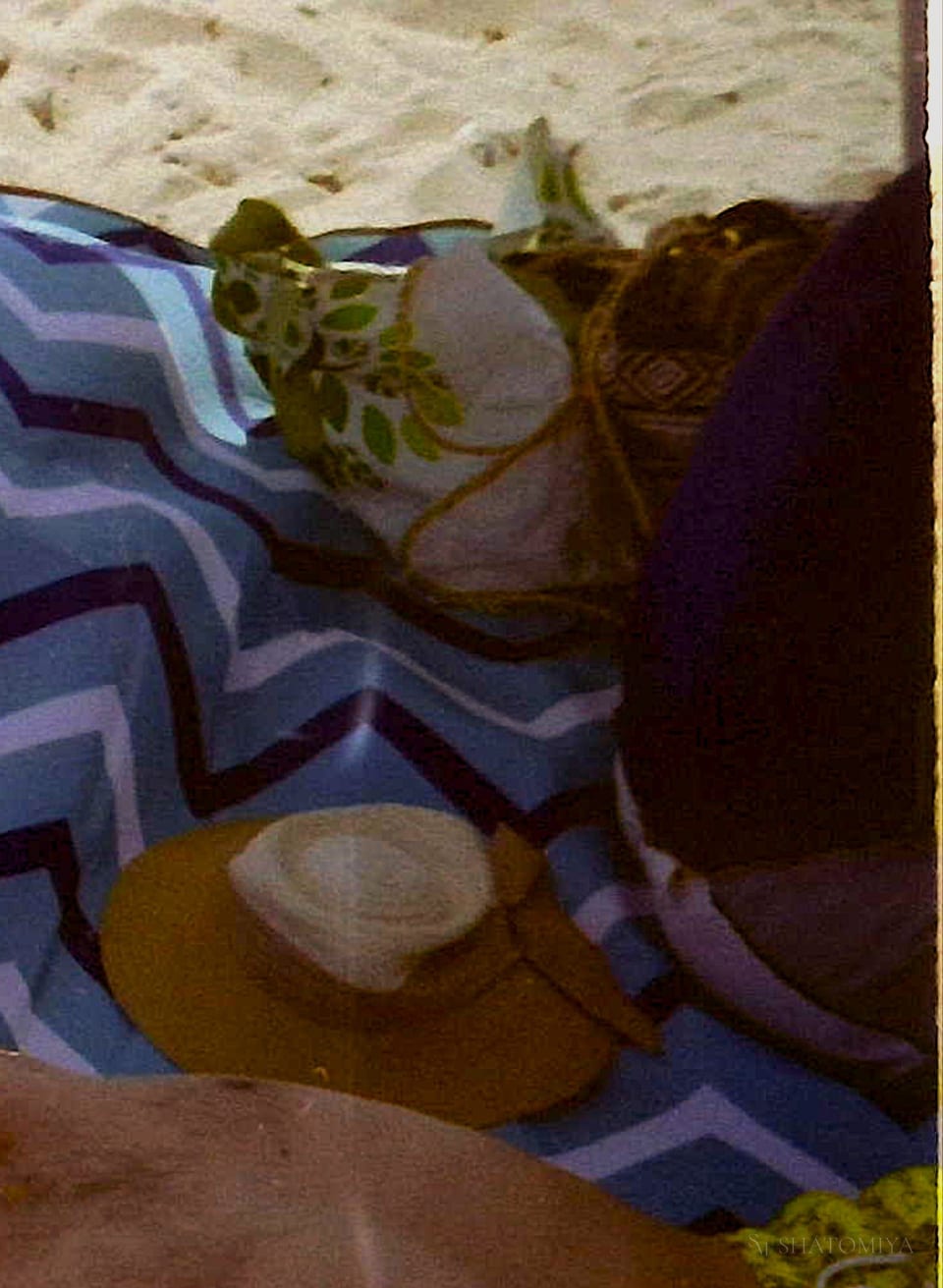 Beach setup with a blue‑and‑white zigzag blanket spread on the sand. On top lie a yellow sunhat, a green leaf‑patterned bag, and a woven geometric bag, arranged neatly for a relaxed day at the beach.